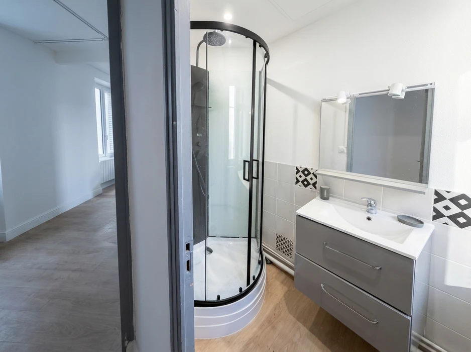 Salle de bain moderne avec cabine de douche quart de cercle à parois en verre et profilés noirs, vasque encastrée sur meuble suspendu gris laqué deux tiroirs, robinetterie chromée, miroir éclairé par spots LED, carrelage mural blanc avec frise ciment noir et blanc motif graphique, sol stratifié imitation parquet