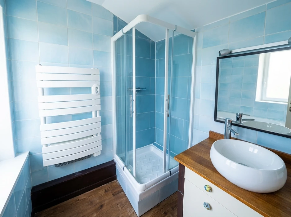 Salle de bains carrelée en bleu clair, avec cabine de douche d'angle à parois coulissantes en verre transparent, cadre blanc, receveur bas relief