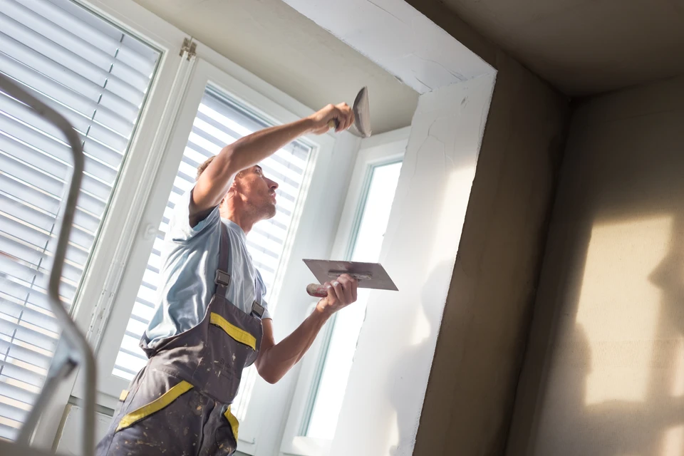 Artisan en salopette applique de l'enduit au couteau sur un mur intérieur près d'une fenêtre avec volets